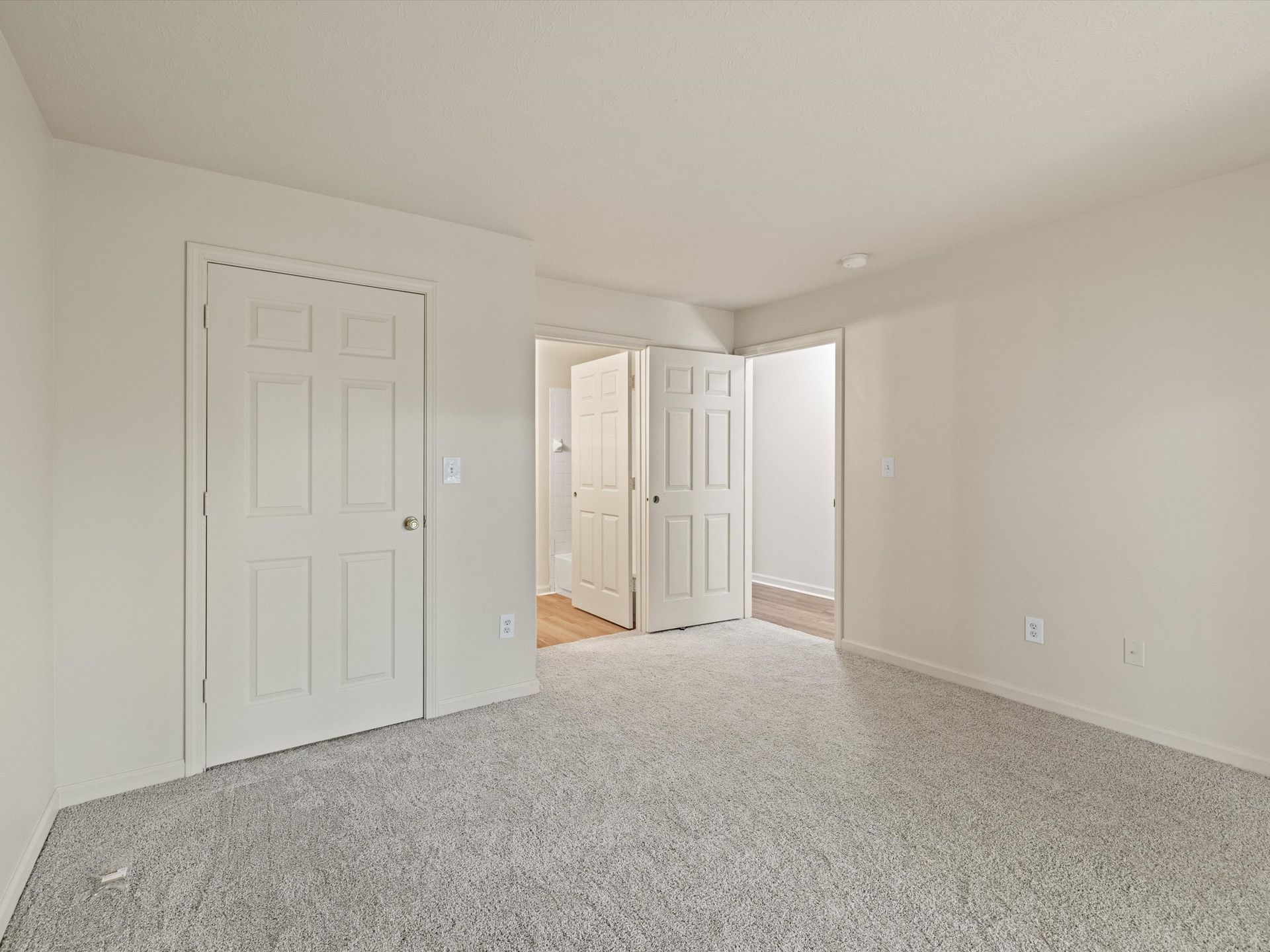 An empty bedroom with a carpeted floor and white walls with view of bathroom and closet door.