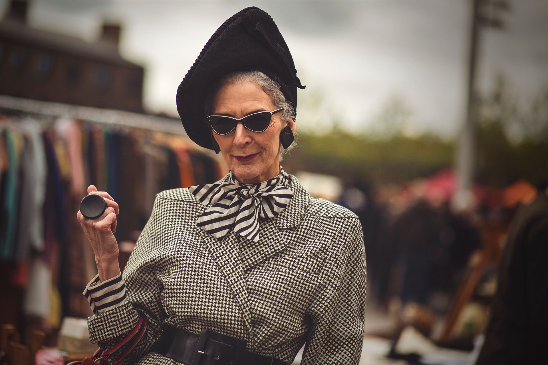 Woman in patterned blouse and sunglasses posing at an outdoor market, holding a small object.