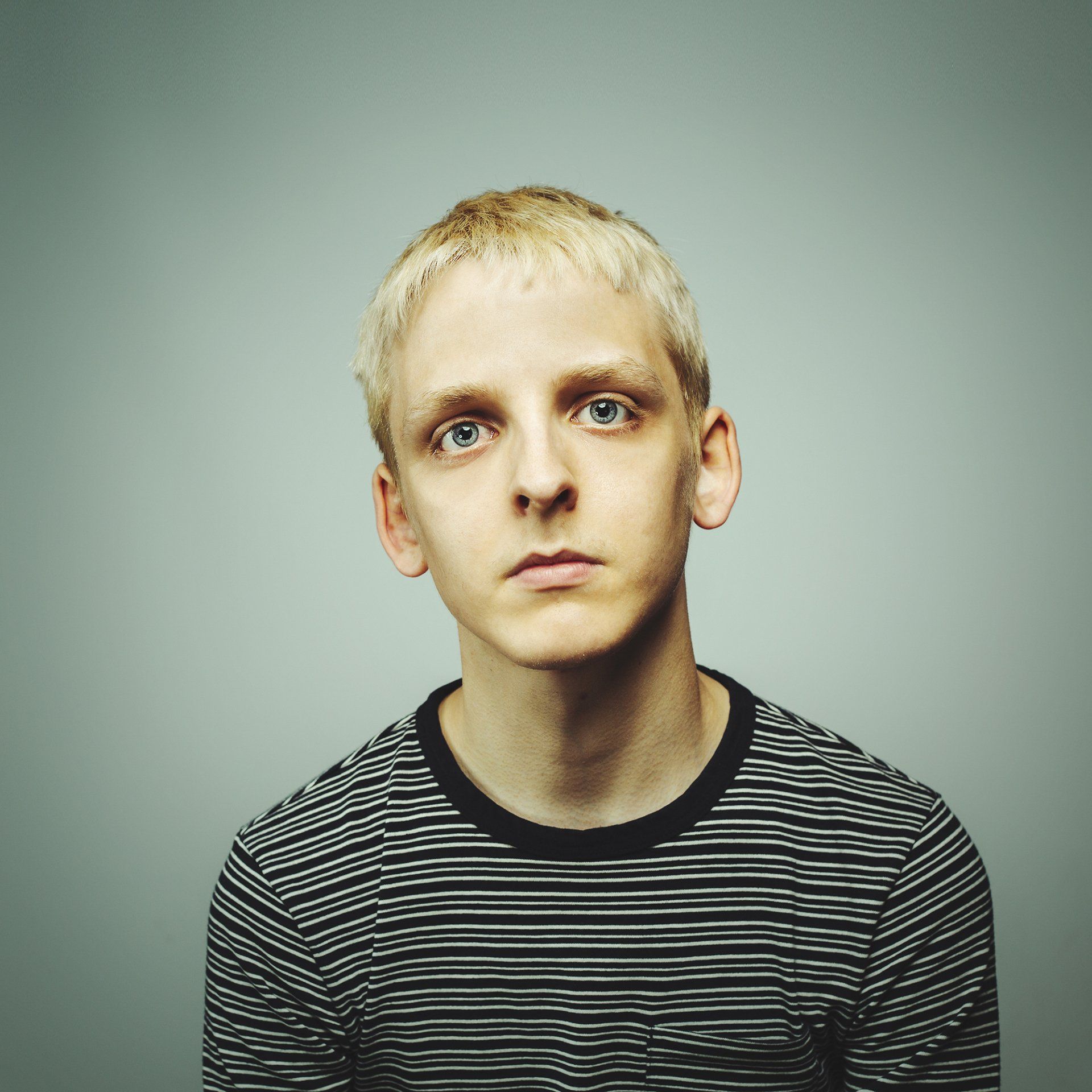 Portrait of a blond person in a striped shirt against a plain gray background, looking at the camera.