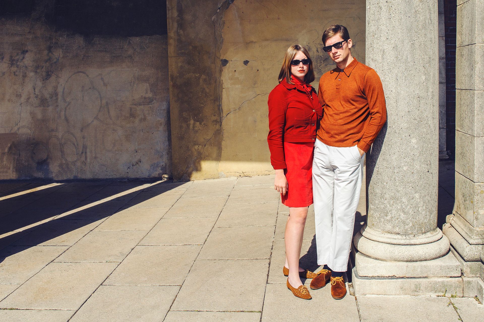 Two people standing by a stone column in bright sunlight, wearing red and orange outfits.