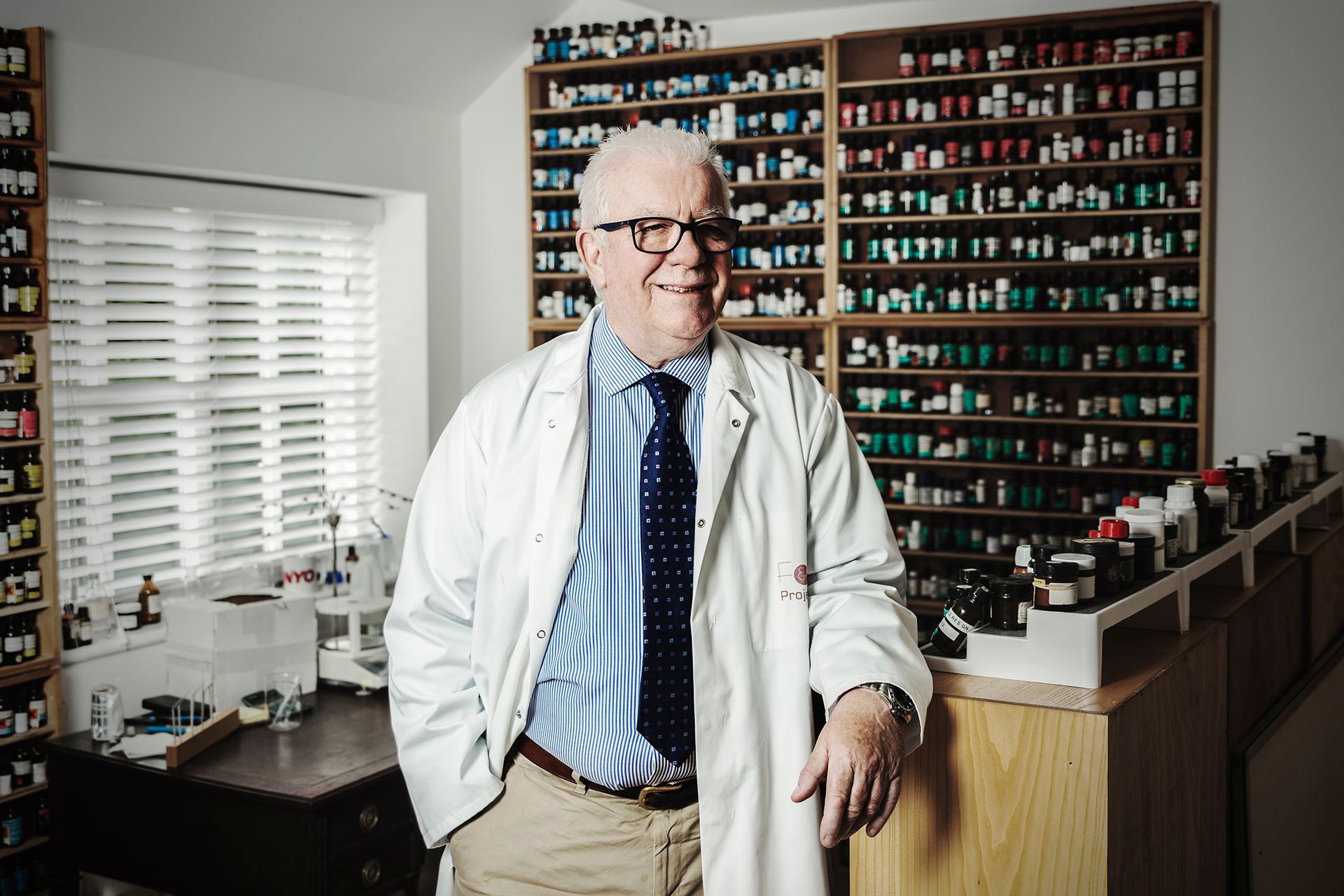 Man in a white coat standing in a lab or office with shelves of small bottles behind him