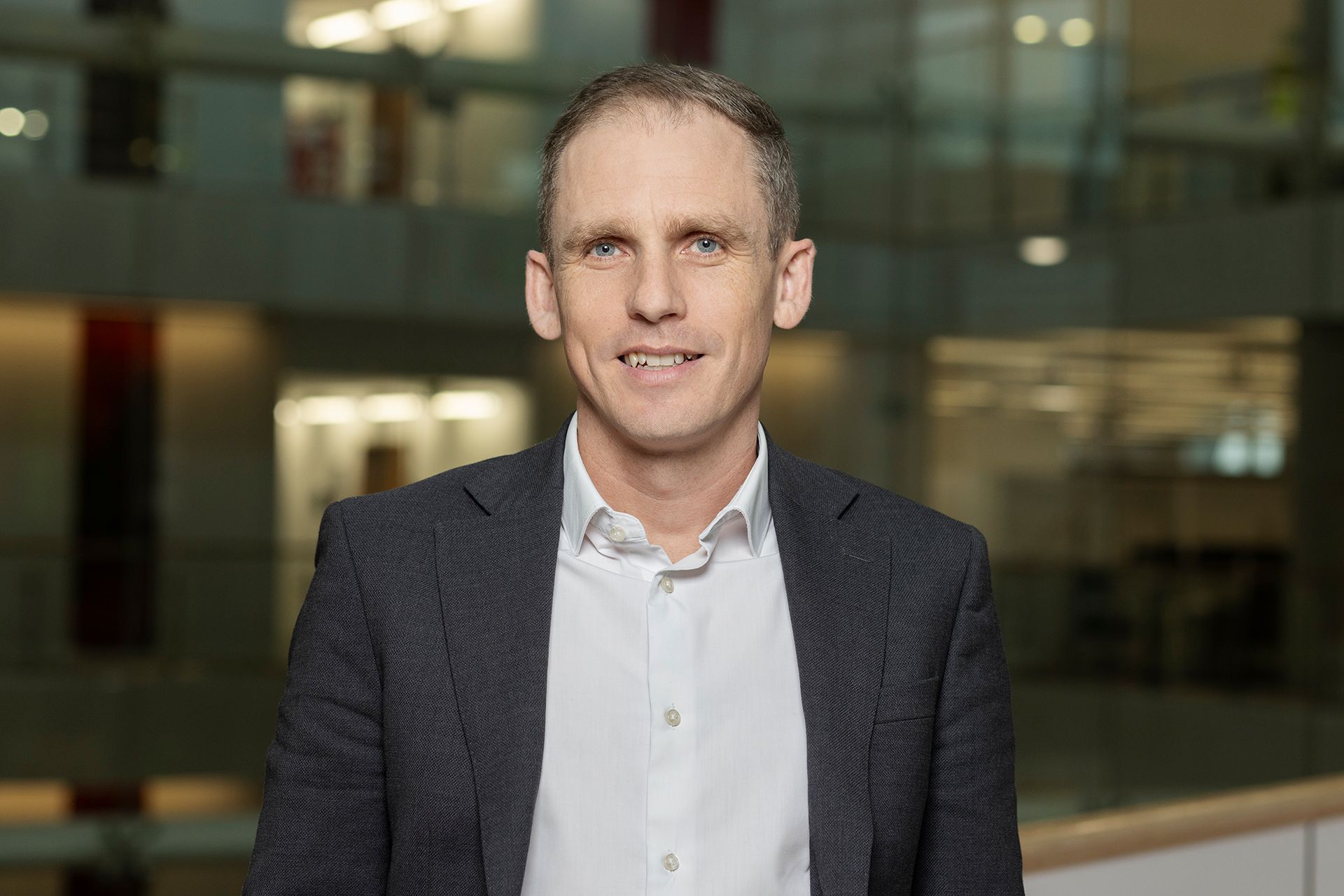 Person in a dark blazer and white shirt standing indoors with a blurred glass office background Portraits & Headshots - Copyright Dammo Photography