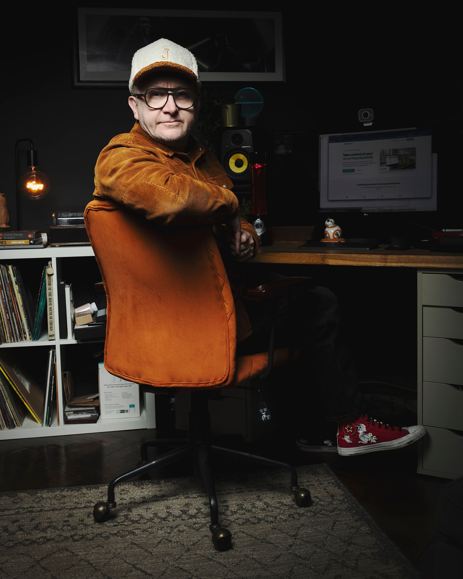 Person in an orange jacket and white cap sitting at a desk in a dim home office.