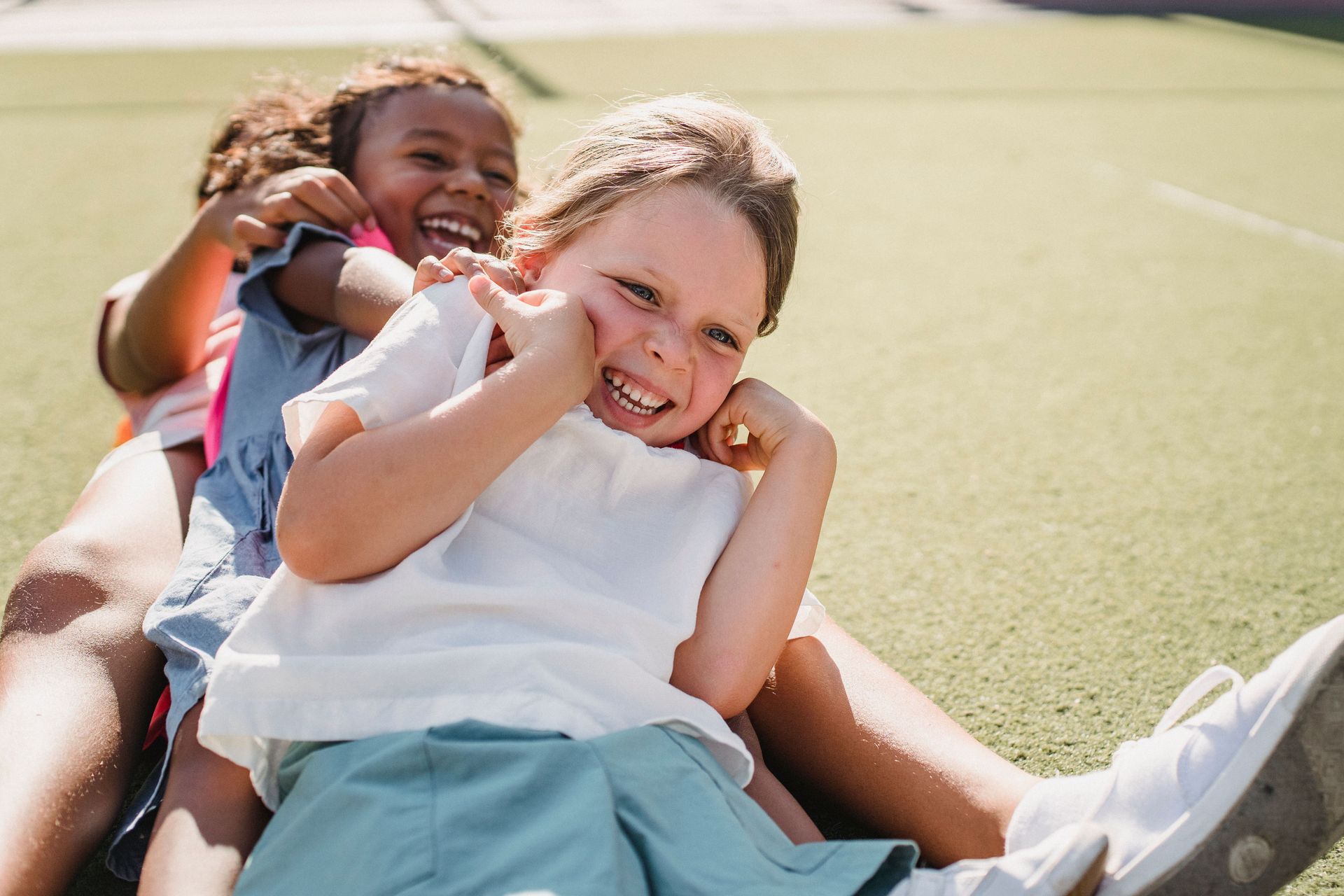 Two children laughing and sliding together on a sports field in bright sunlight