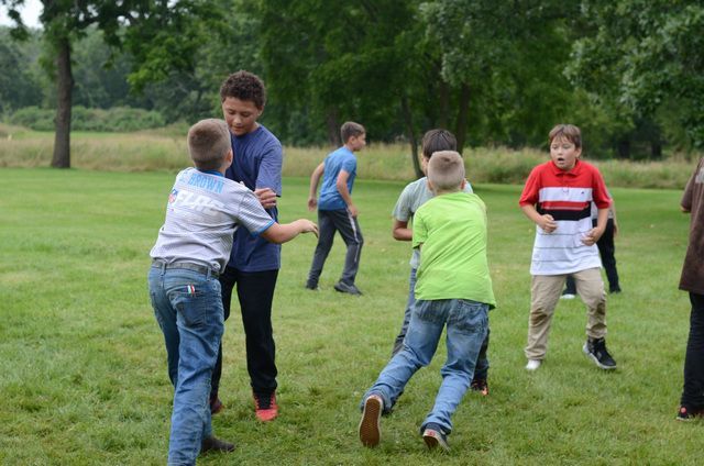 Children playing tag on a grassy field outdoors, with one child in blue and another in green wrestling playfully.