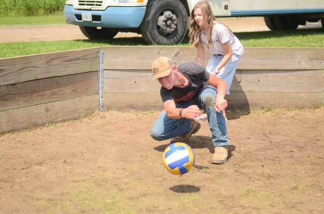 Two people play with a blue-and-yellow ball on dirt near a wooden barrier outdoors.