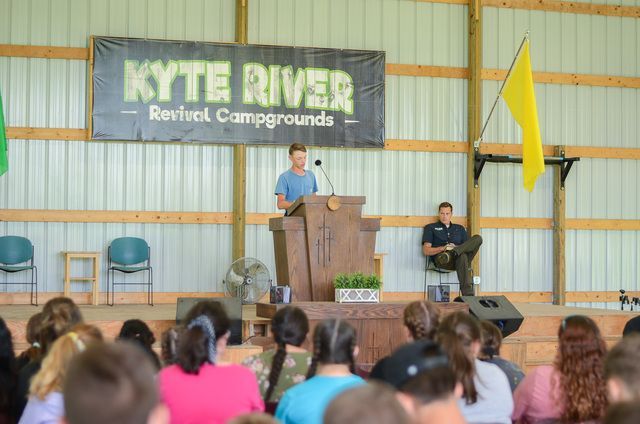 Speaker at podium on stage at Kute River Revival Campgrounds with seated audience and yellow flags