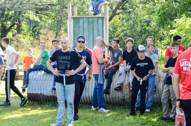 Group of people gathered outdoors by a large tire obstacle, some holding poles and preparing for an activity