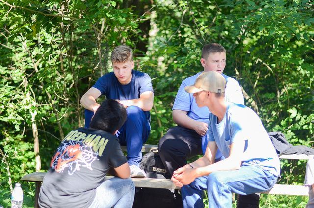Four young people sit and talk at an outdoor picnic table in a sunny wooded area.