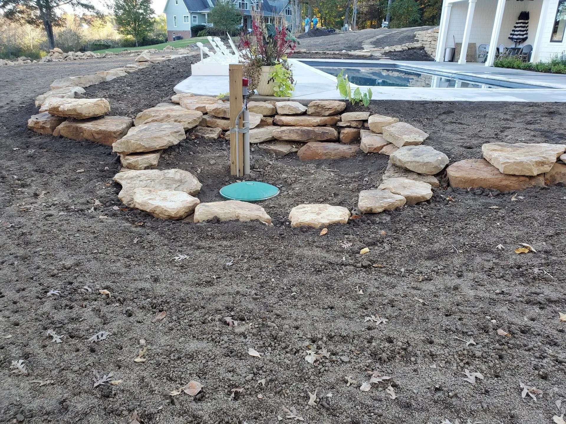 Stone-bordered planting bed with a young tree and green mulch in a landscaped yard beside a patio