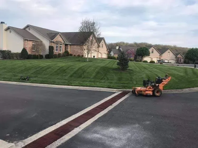 Orange lawn mower on a paved street beside a grassy hill and large houses at dusk