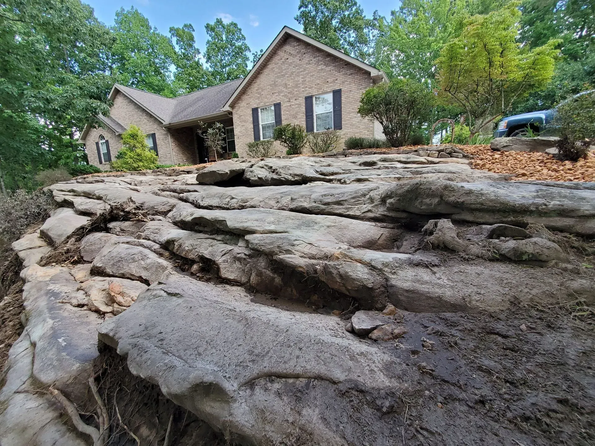 Stone retaining wall in front of a brick house with trees and a car in the driveway.