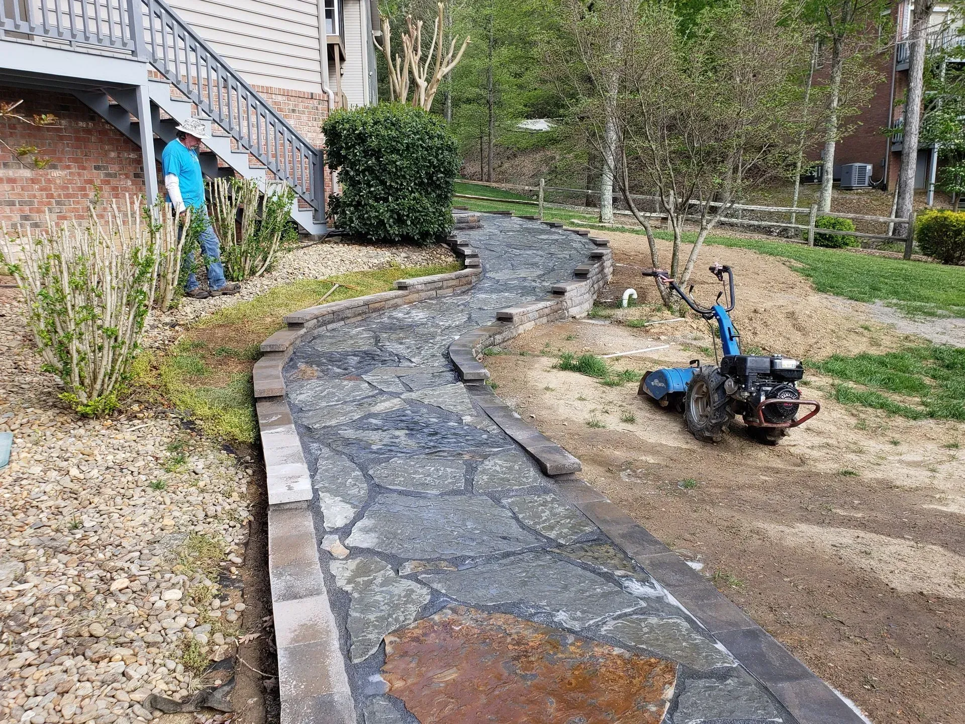 Stone walkway curves through a yard beside a house, with a blue wheelbarrow on the dirt beside it.