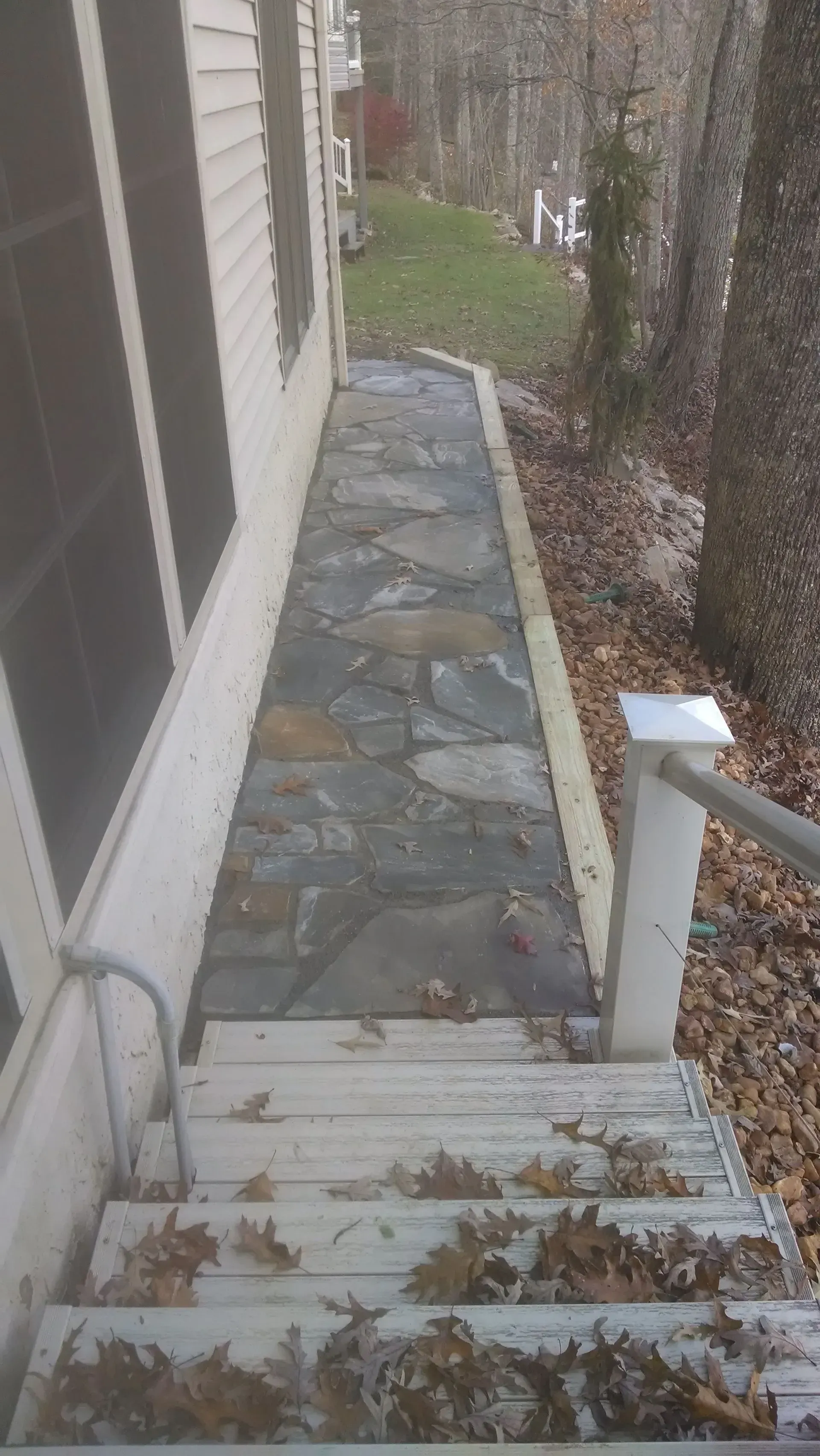 Narrow stone walkway beside a house, viewed from a small set of stairs, with fallen leaves and shrubs.