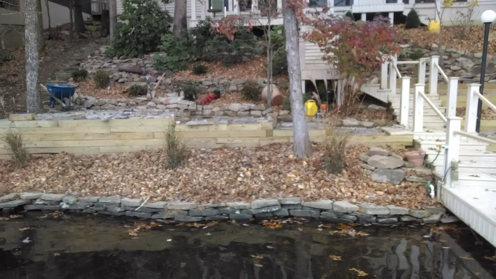 Rocky waterfront garden with a wooden dock, stairs, and autumn trees behind a low stone wall