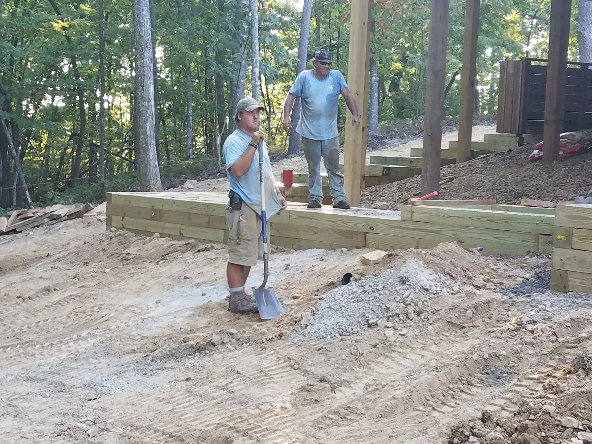 Two workers on a wooded construction site, one holding a shovel near a dirt mound
