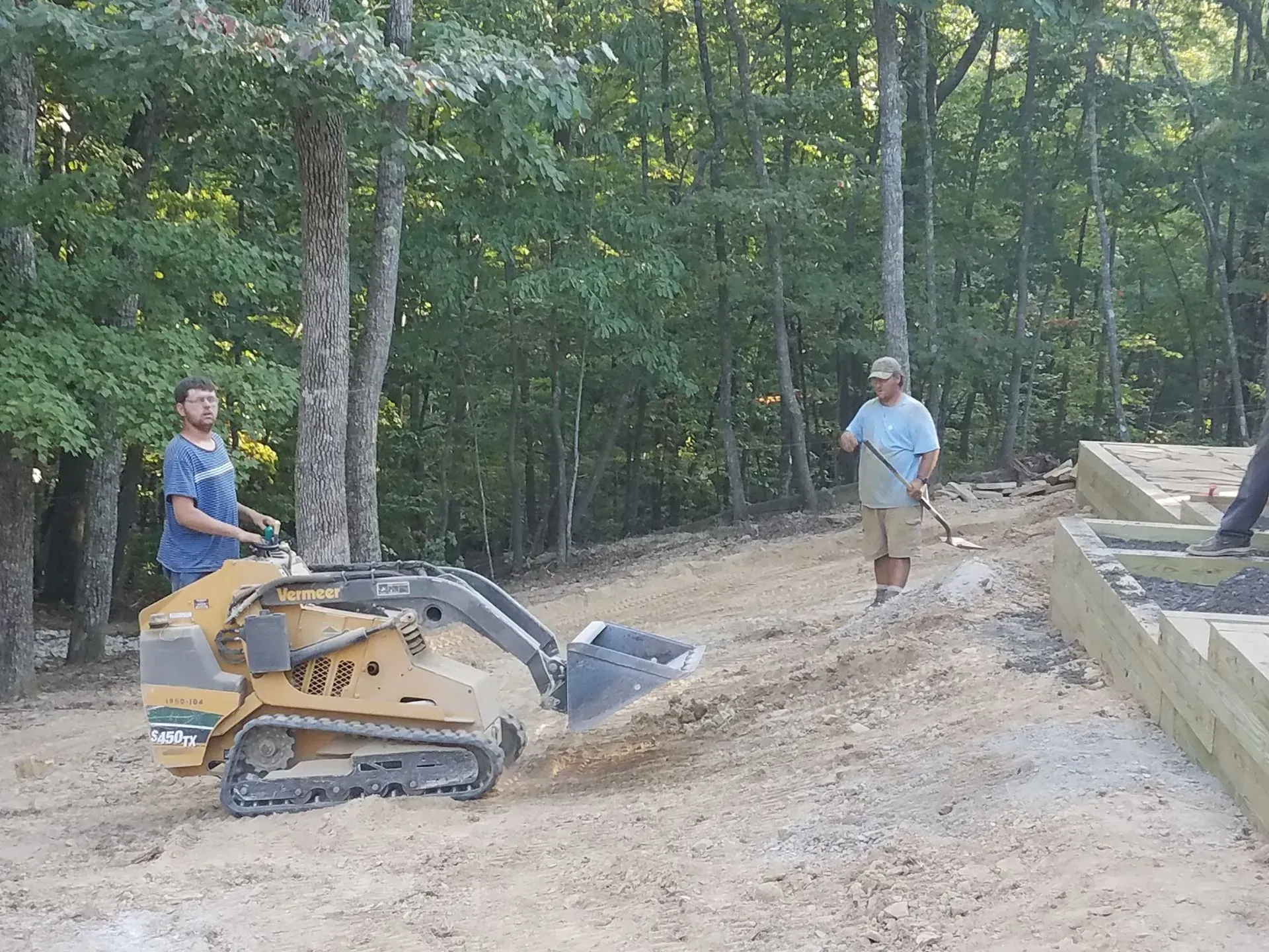 Workers operate a tracked machine on a dirt path beside a stone retaining wall in a wooded area.