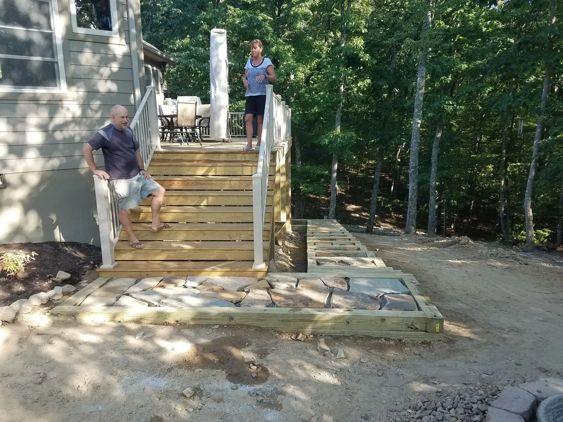 Two people on outdoor wooden stairs beside a house, with a stone path and trees in the background.