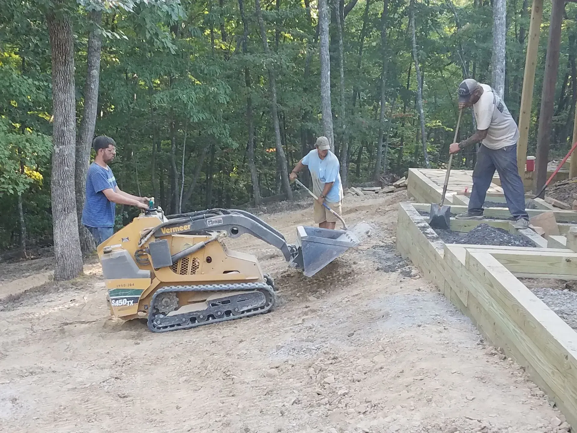 Three workers operating a tracked loader and building a retaining wall in a wooded outdoor site