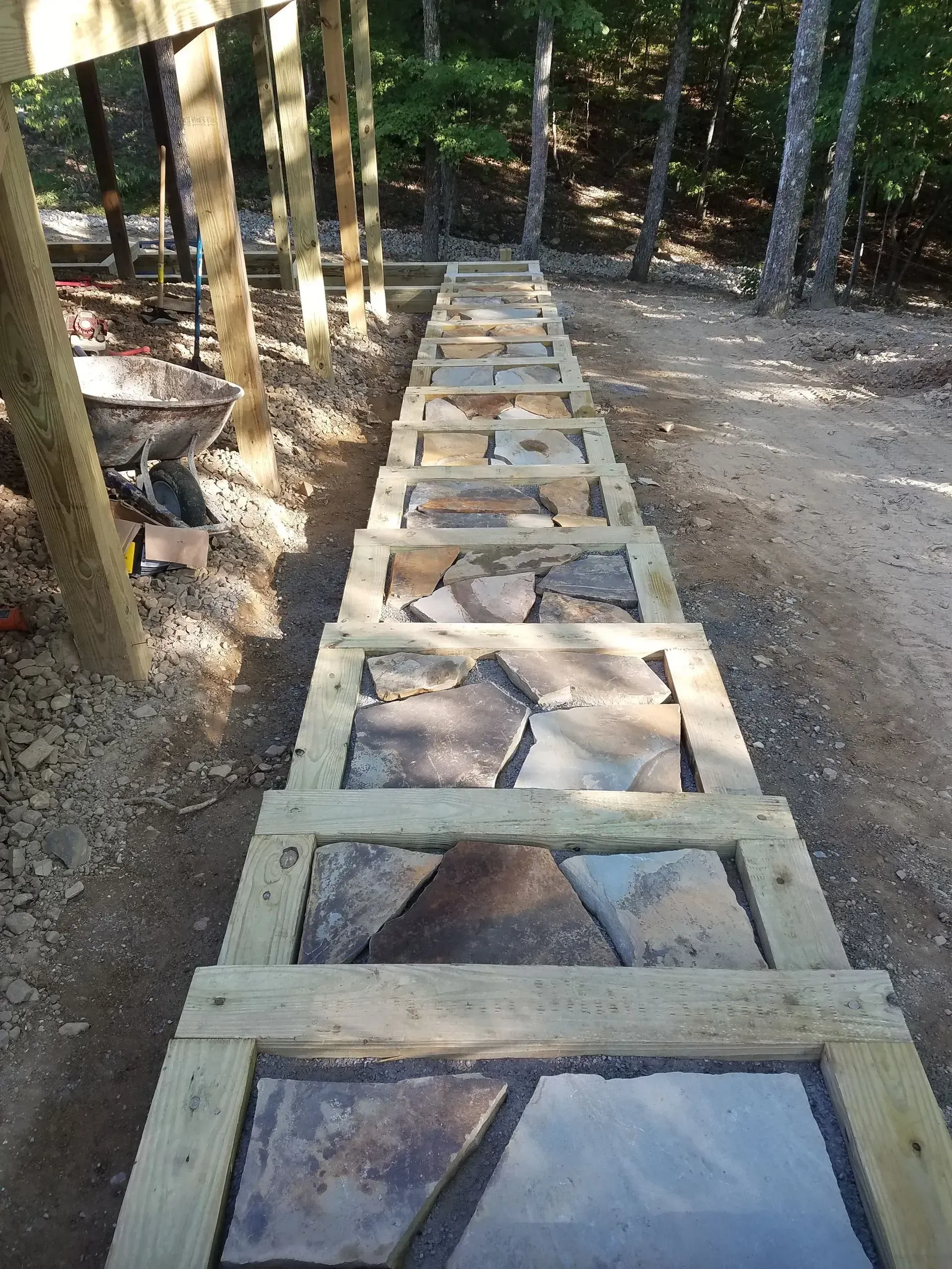 Wooden boardwalk under construction in a wooded area, with concrete footings and tree shadows.