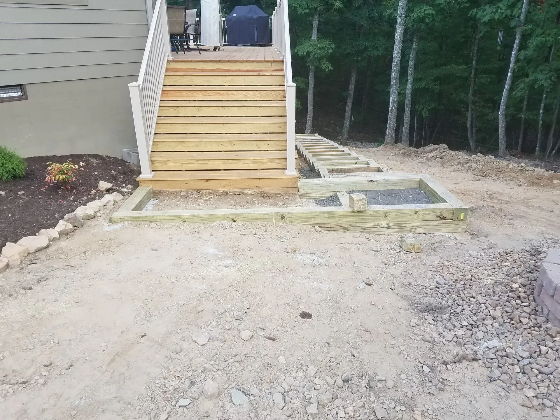 Gravel yard with wooden steps leading to a side entrance and a small concrete pad beside a wooded area