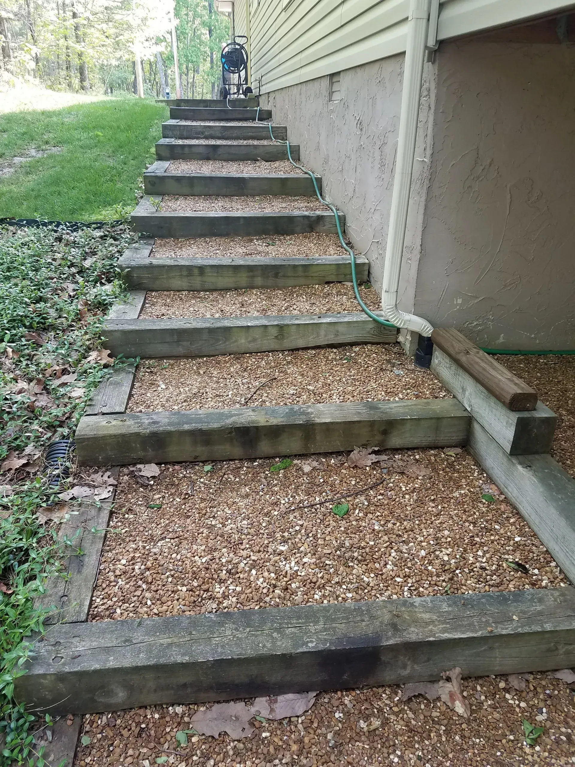Steep outdoor wooden stairs with gravel treads beside a house and grassy slope
