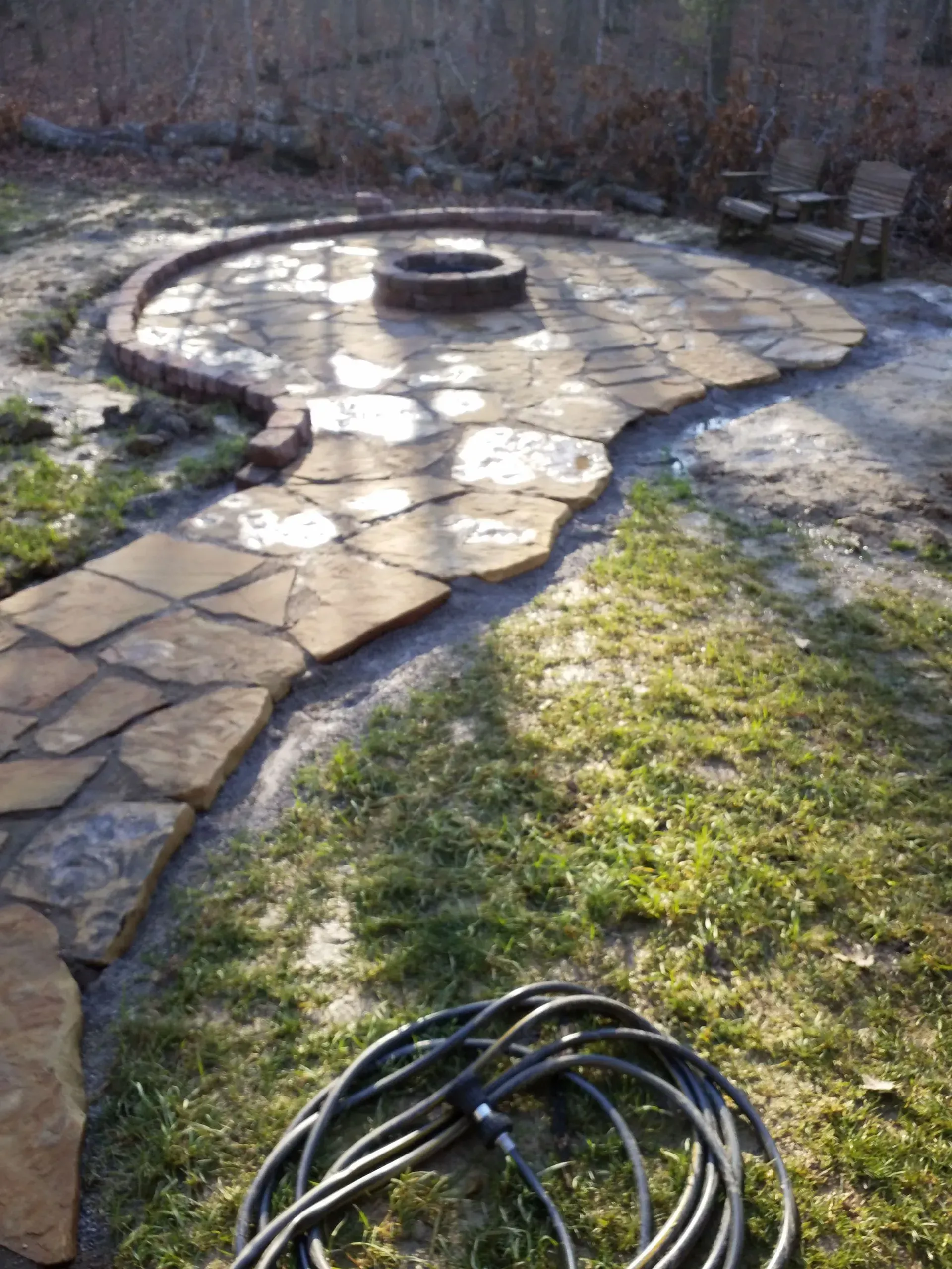 Stone patio with a circular fire pit, bordered by grass and a coiled hose in the foreground.