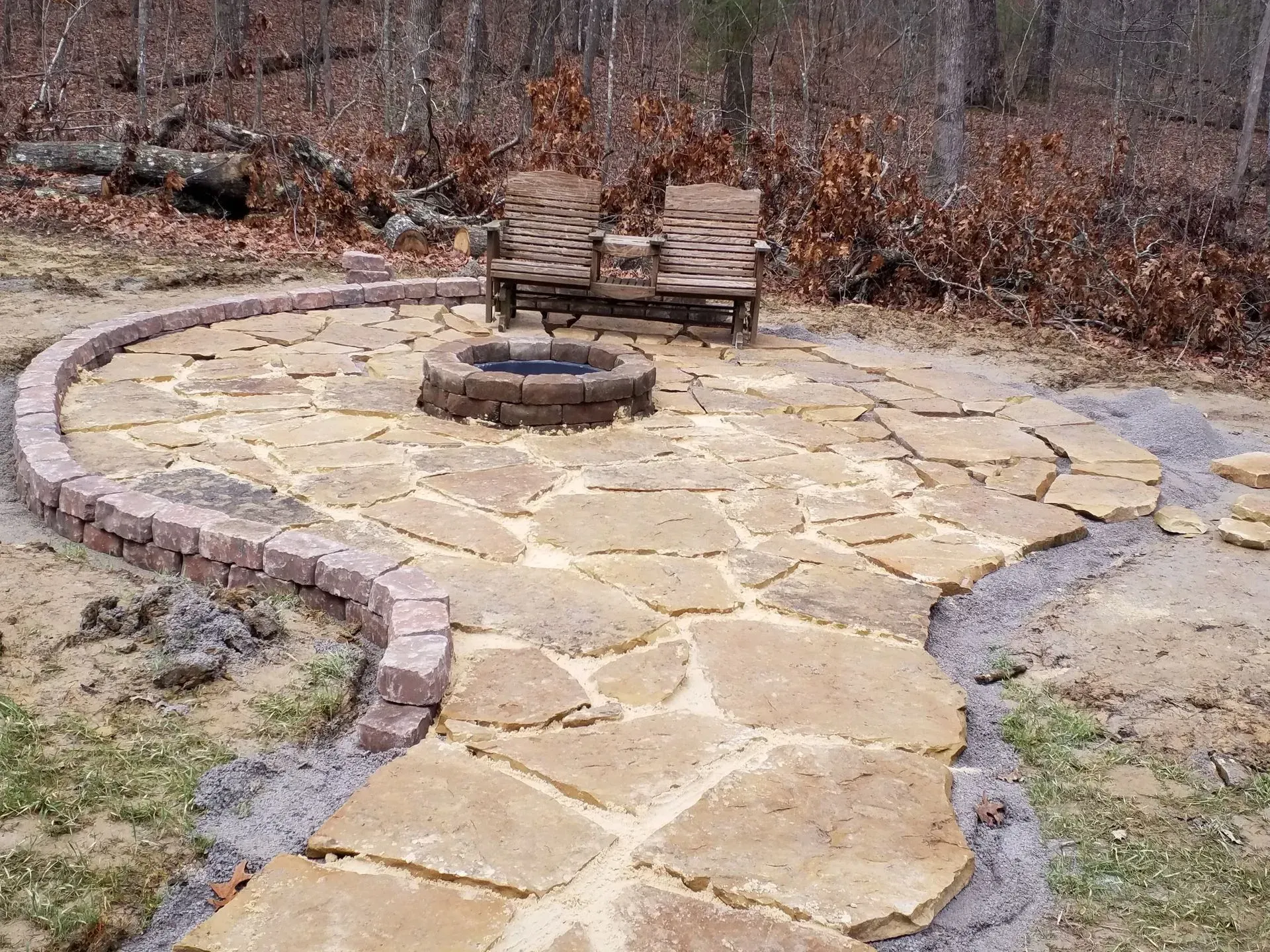 Stone patio with a circular fire pit and two wooden chairs in a wooded backyard.