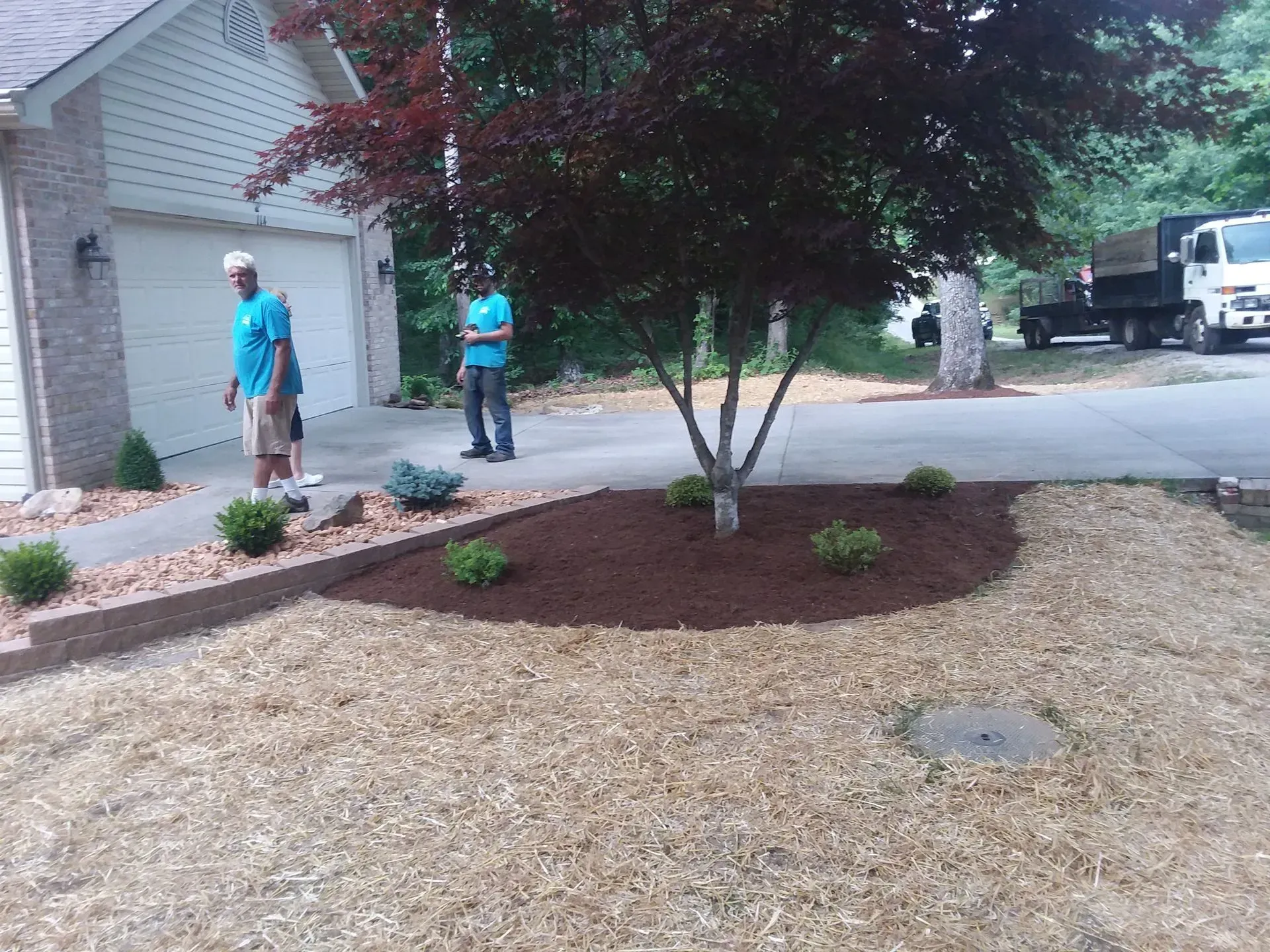Two workers in teal shirts landscaping a front yard with mulch and newly planted shrubs.