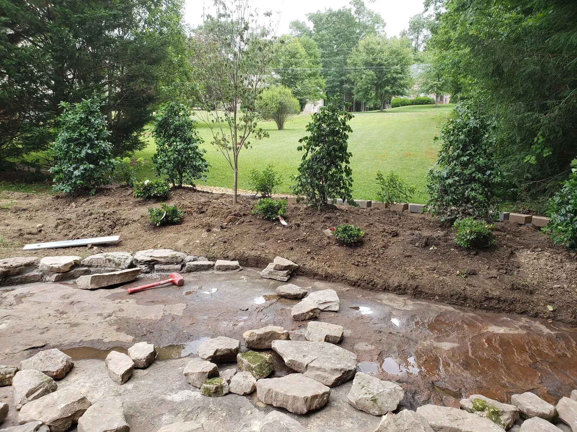 Rock-lined garden stream with shallow water, surrounded by green trees and a grassy lawn