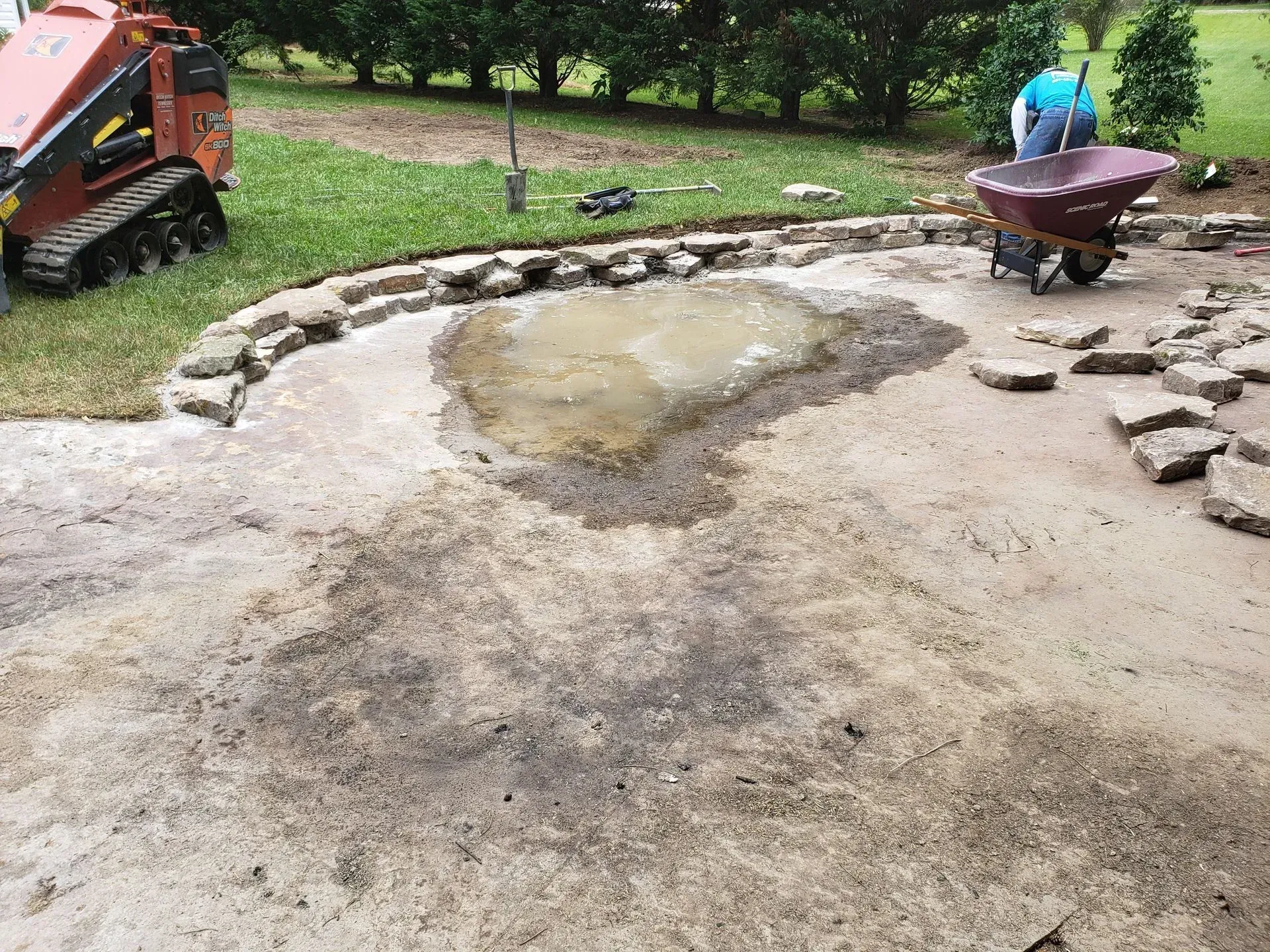 Backyard construction site with a muddy puddle, stone border, and workers near a wheelbarrow and excavator