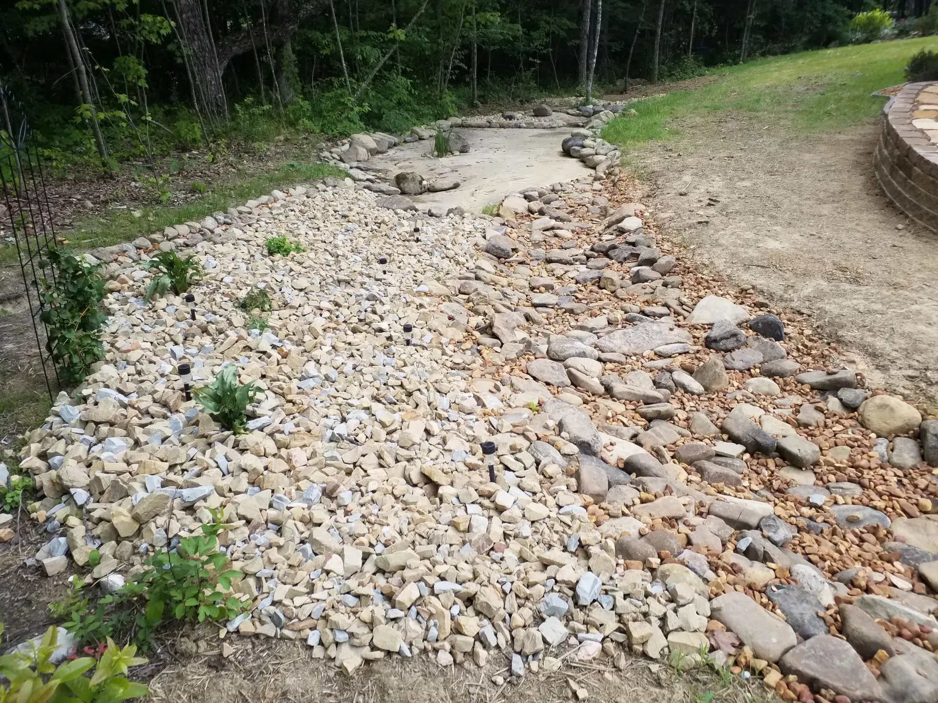 Dry rocky drainage swale beside a grassy yard and wooded edge, with a wooden deck on the right.