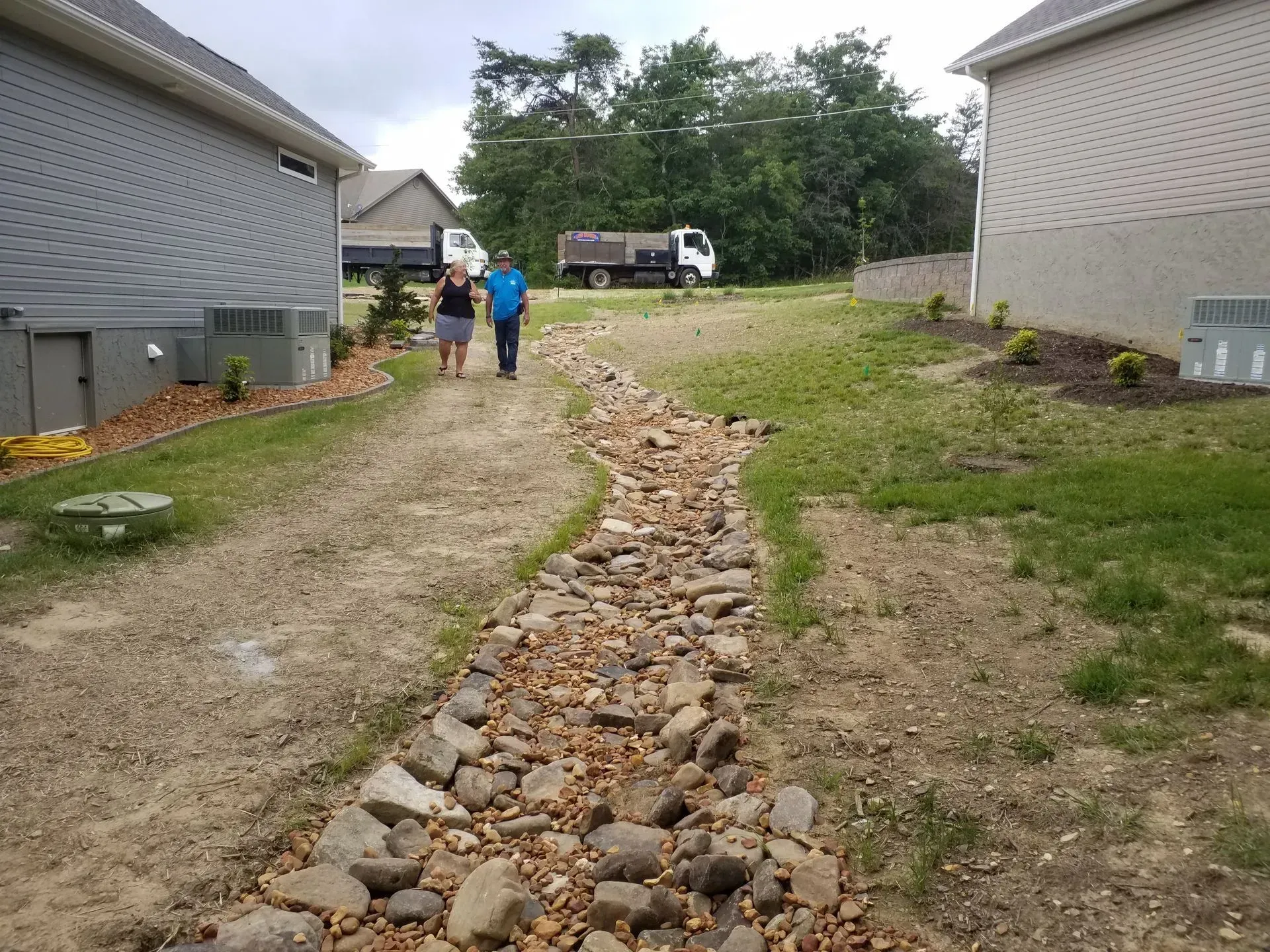 Rock-lined drainage ditch between two houses, with two people walking near a parked truck.