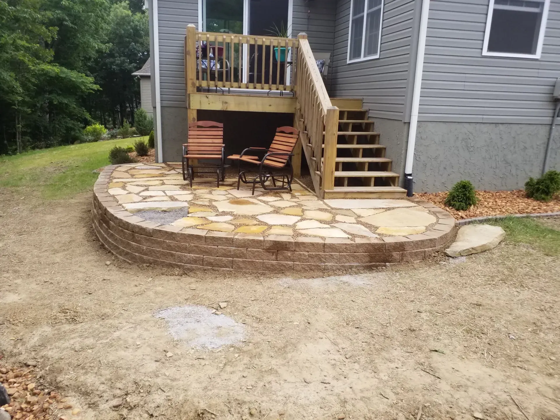 Stone patio with wooden deck stairs and chairs in a backyard