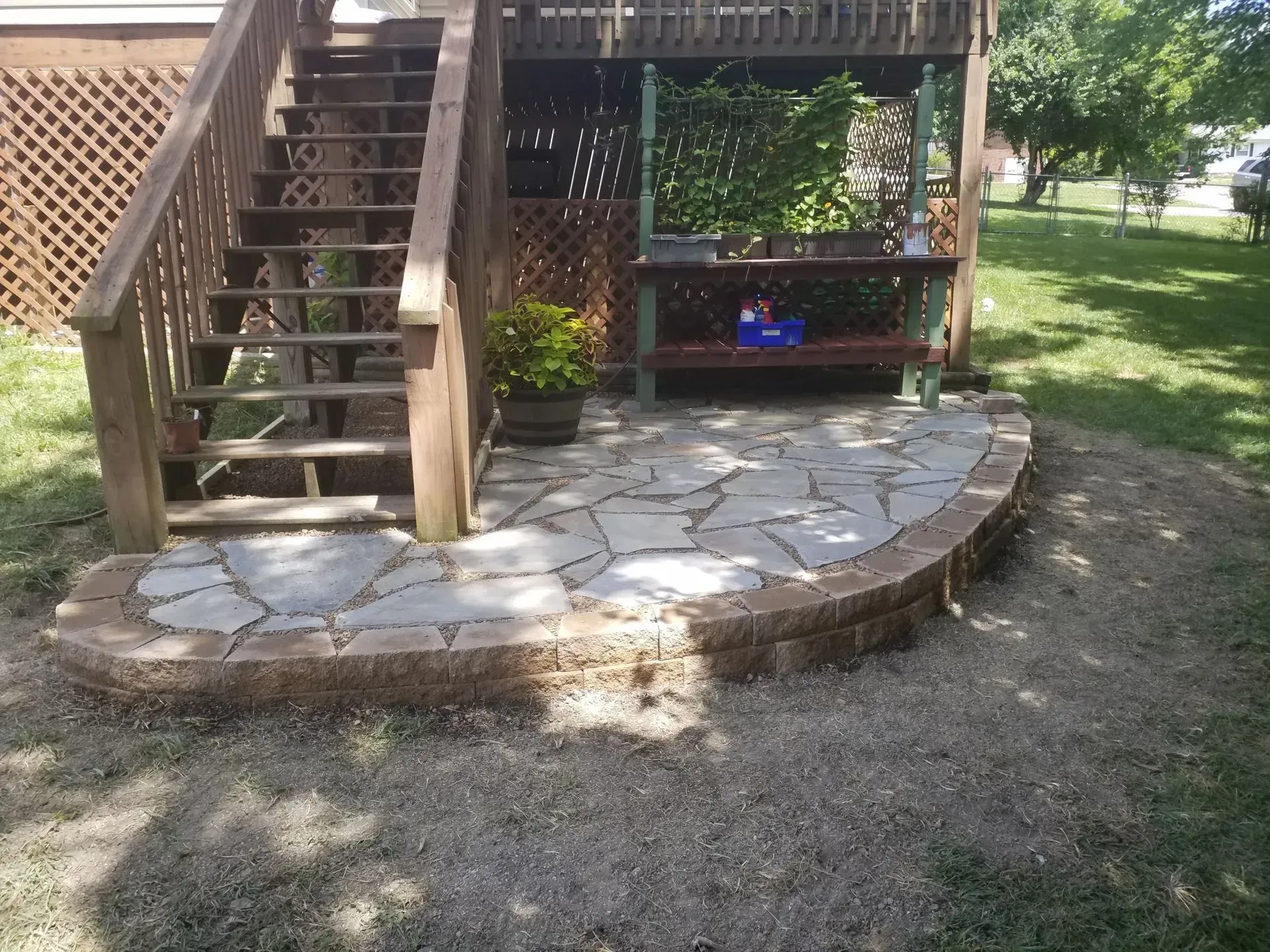 Wooden deck stairs beside a curved stone patio and grassy yard, with potted plants underneath.