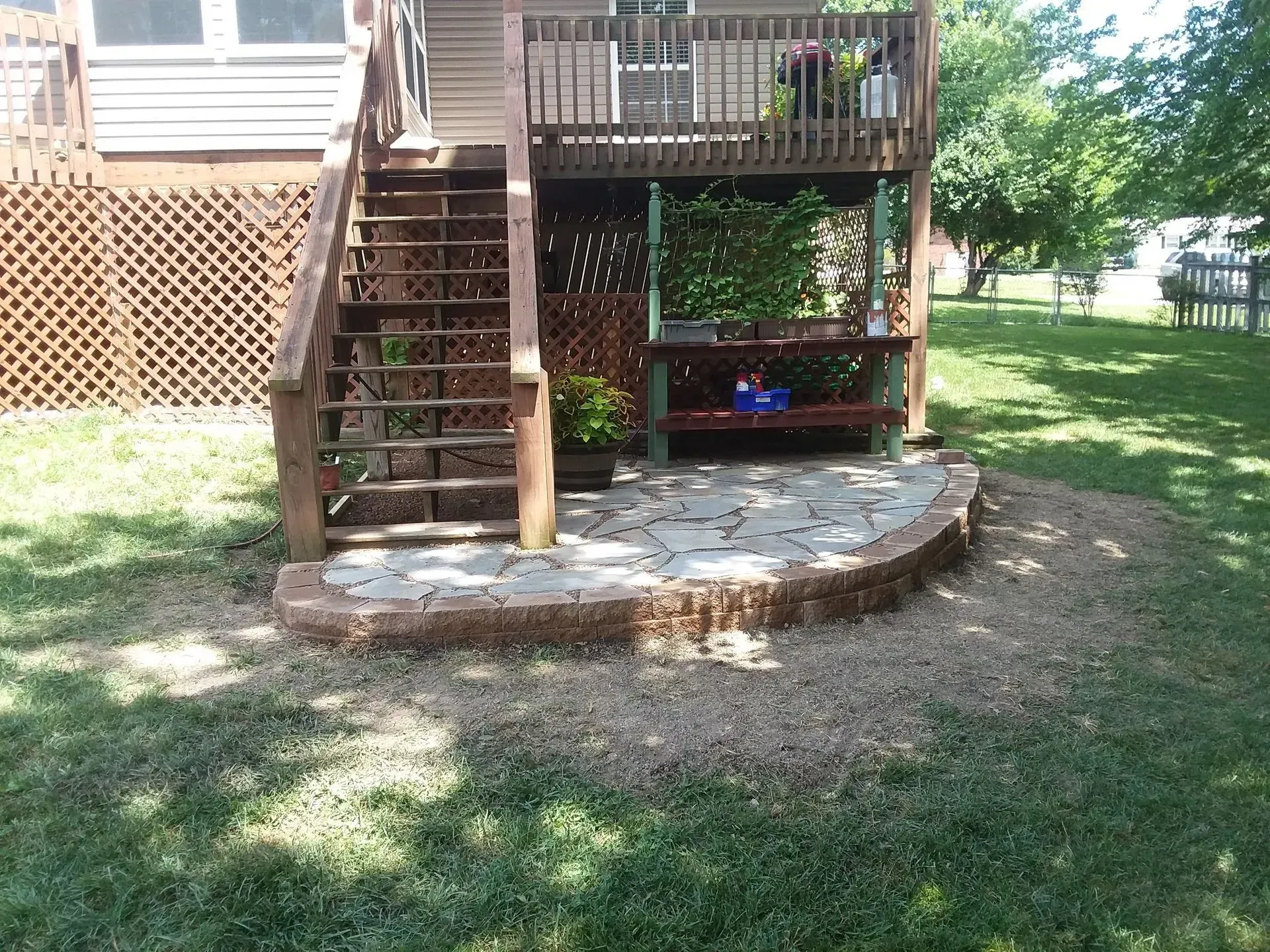 Backyard deck with wooden stairs, stone patio, and grassy yard in sunlight