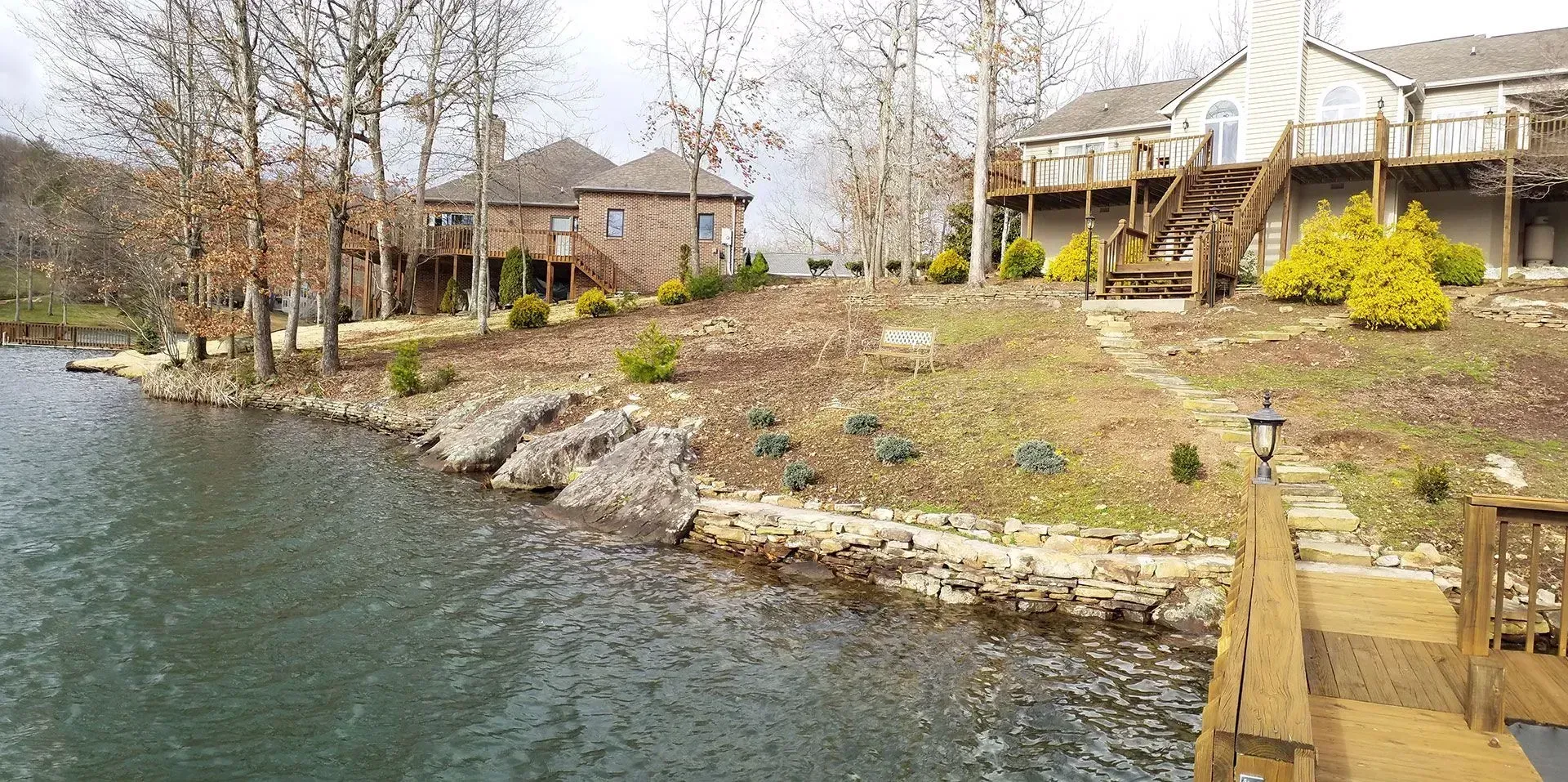 Rocky lakeside shoreline with docks, houses, and spring trees under a cloudy sky