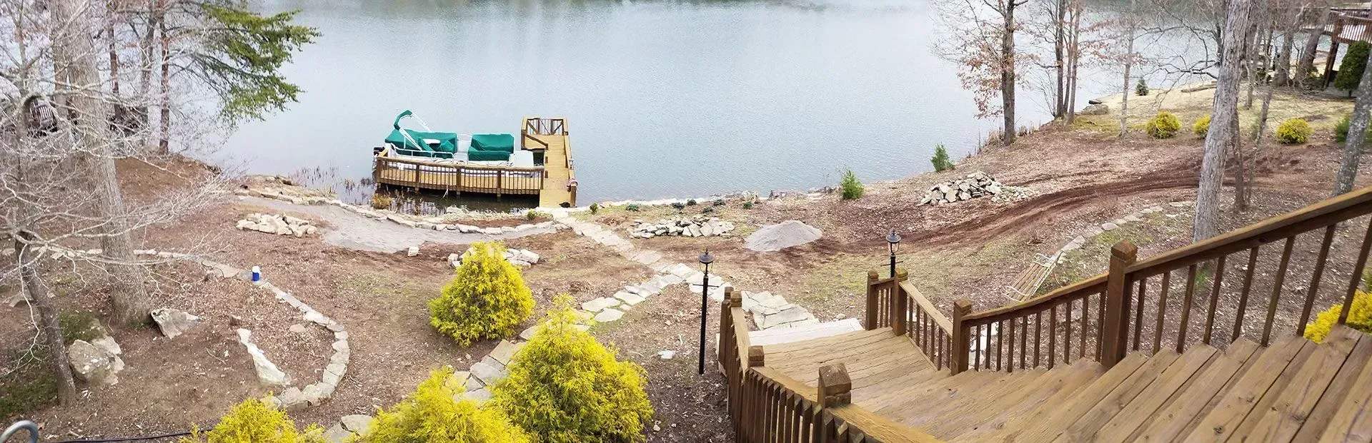 Wooden steps descend to a lakeside dock with a covered boat on a calm, tree-lined shore