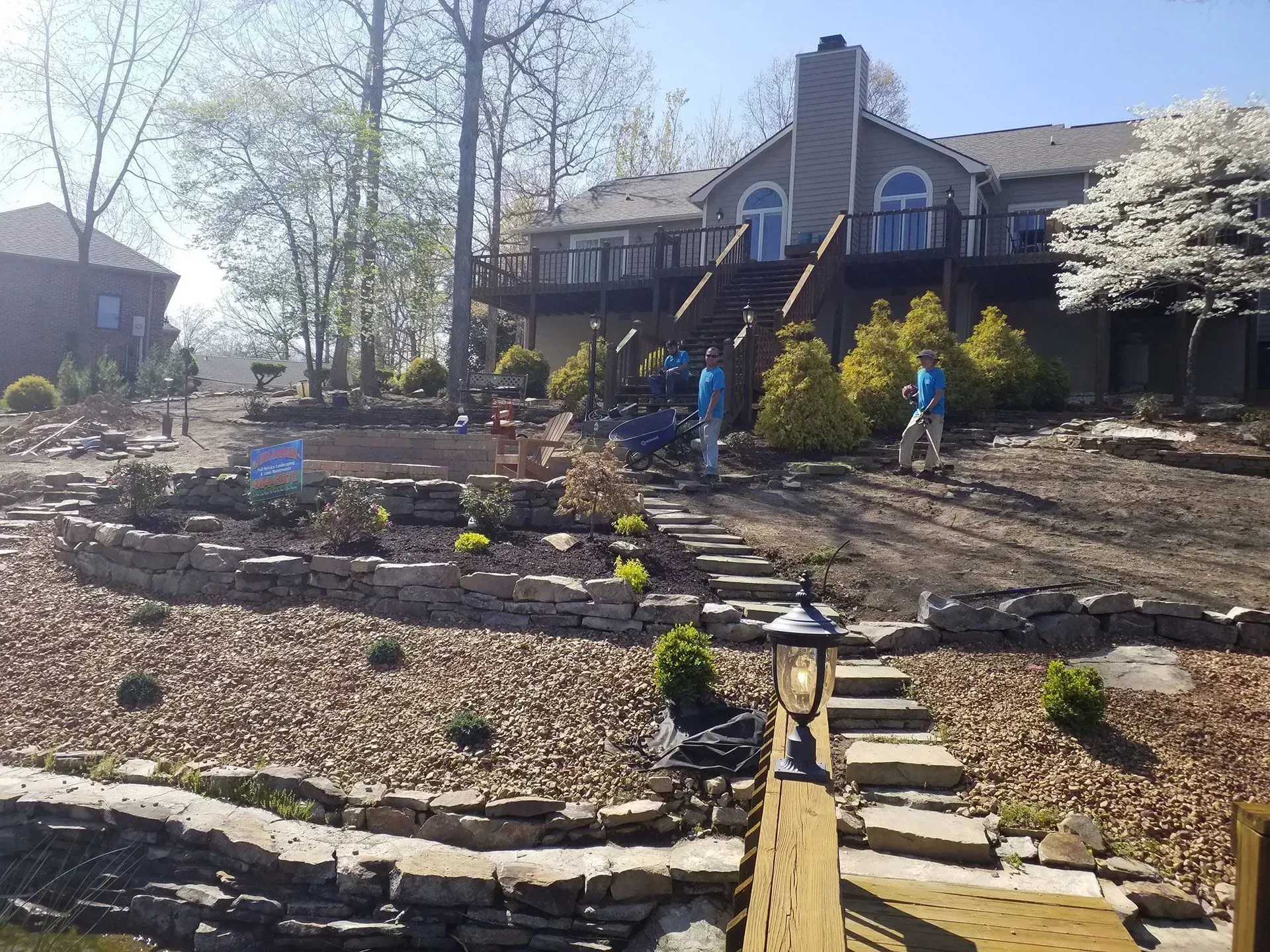 House with stone terraced front yard, steps, and two people in a sunny garden.