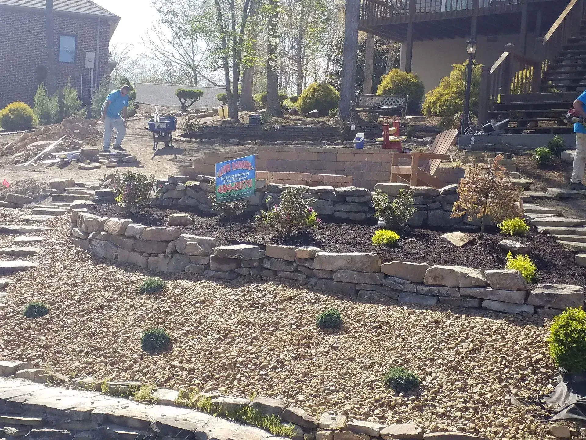 Rock garden landscape with stacked stone borders, a colorful sign, and people working in the background