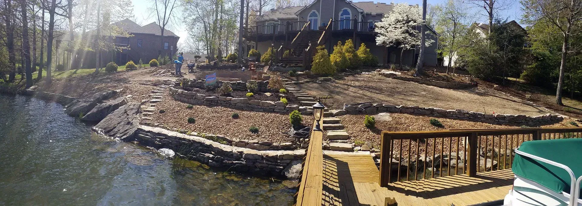 Rocky riverside terrace with stone steps, wooden walkway, and houses among trees in bright sunlight