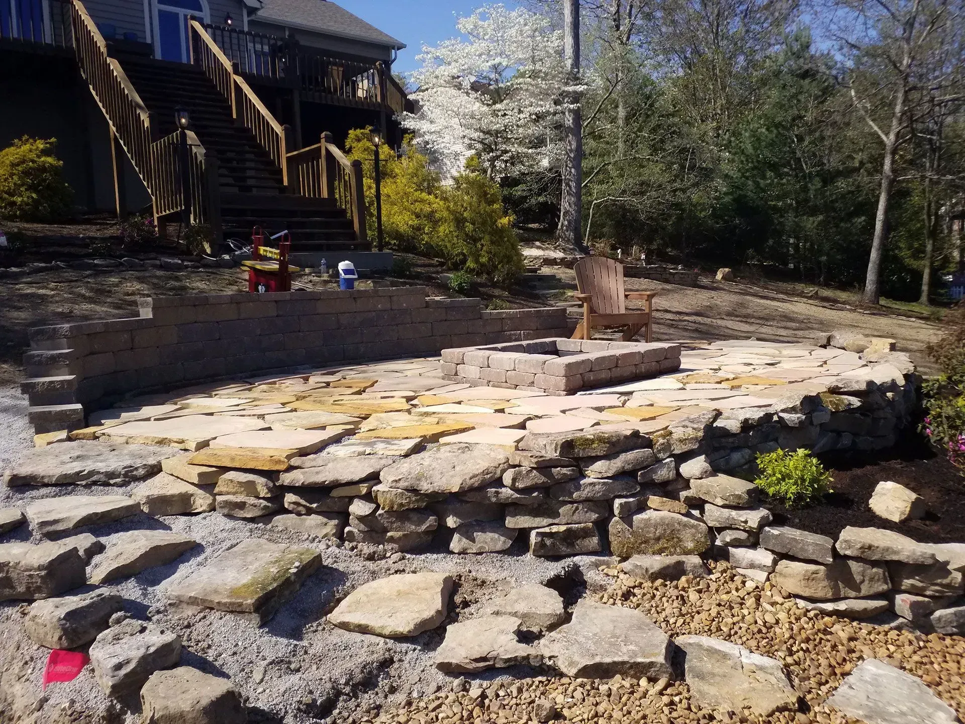 Stone patio and fire pit in a yard, with a house, stairs, and blooming trees in the background.