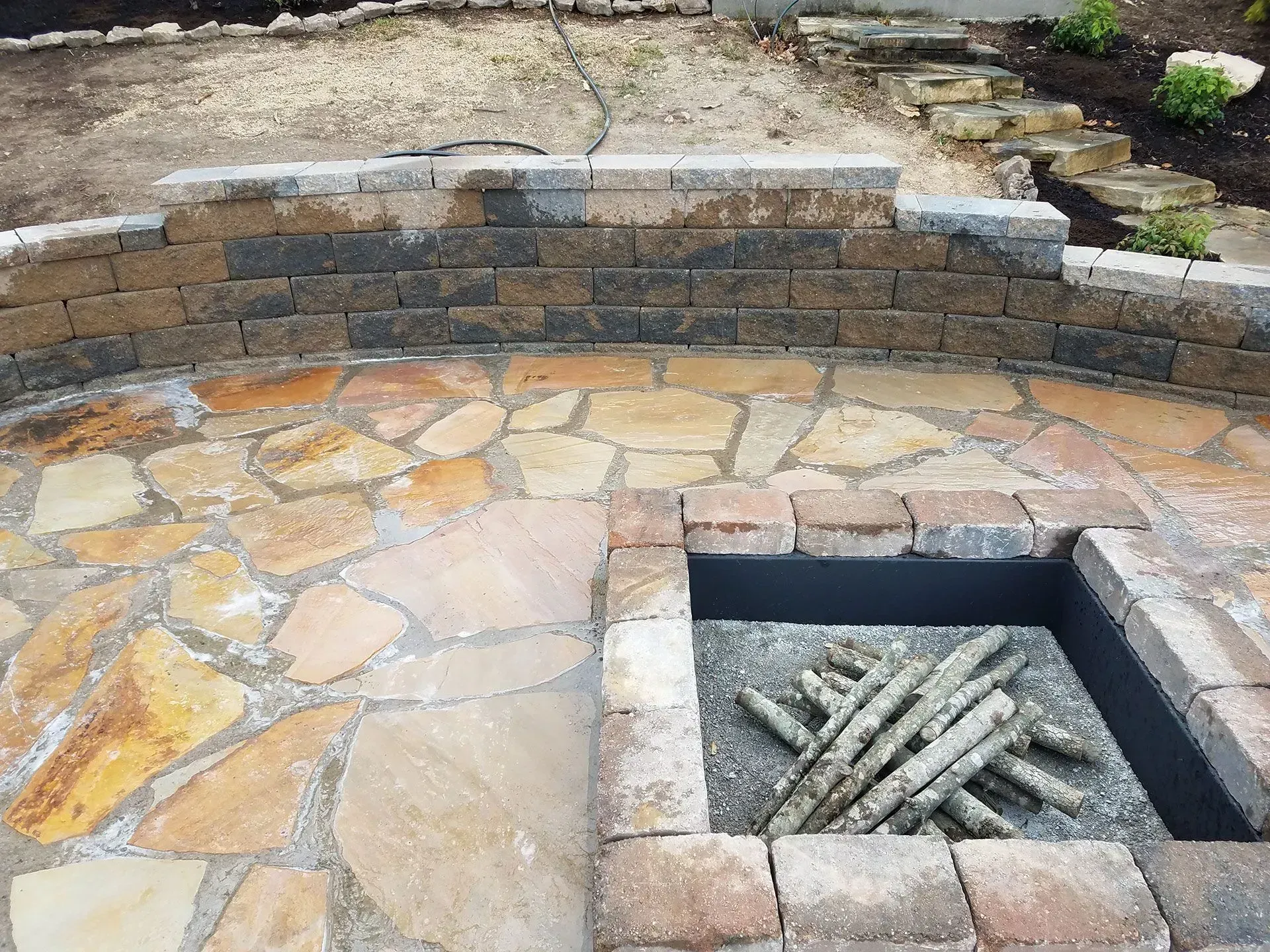 Stone patio with a square fire pit containing stacked logs, bordered by a curved stone wall.