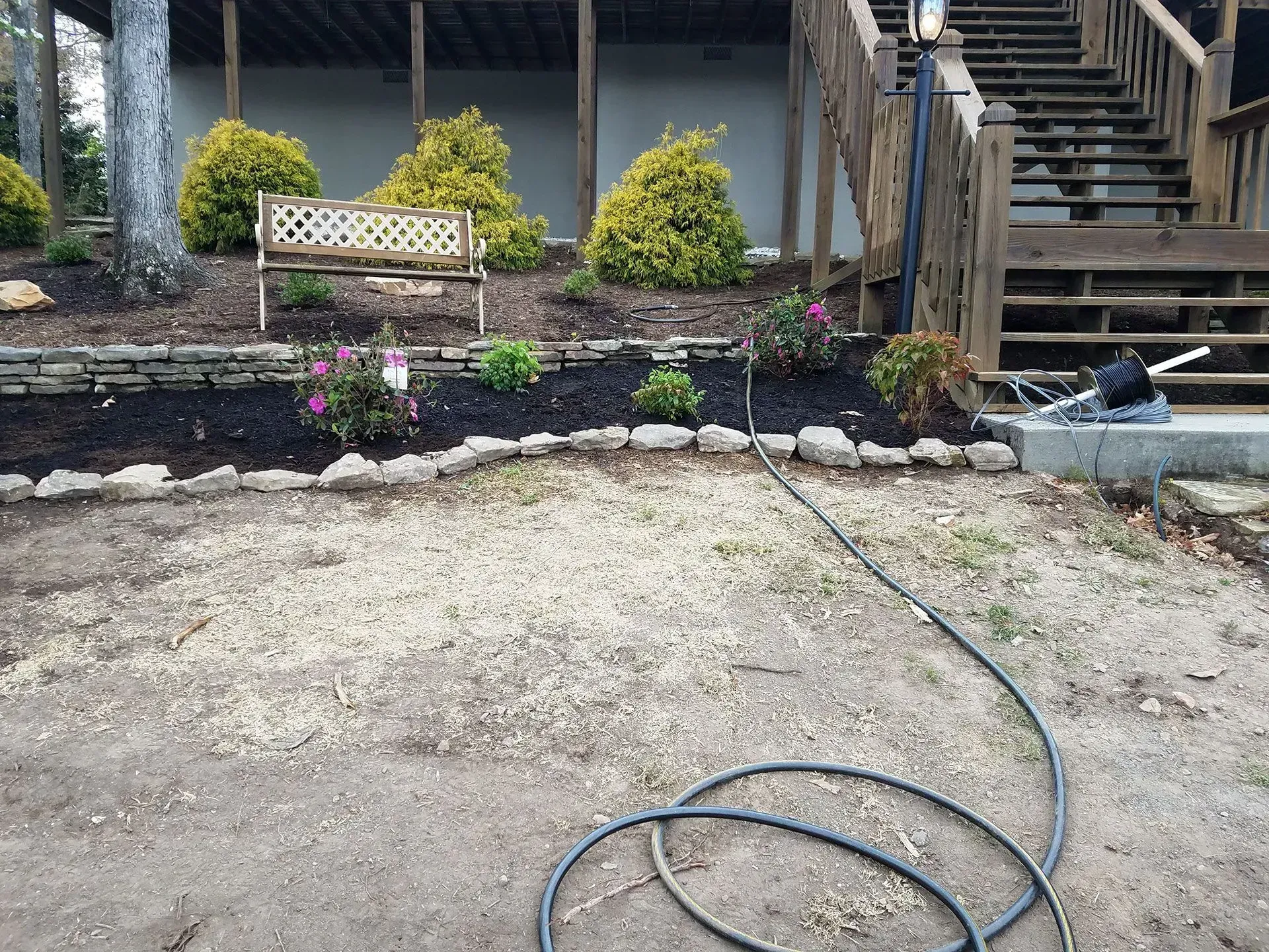 Backyard garden bed with shrubs, a bench, stone border, stairs, and a hose on bare ground