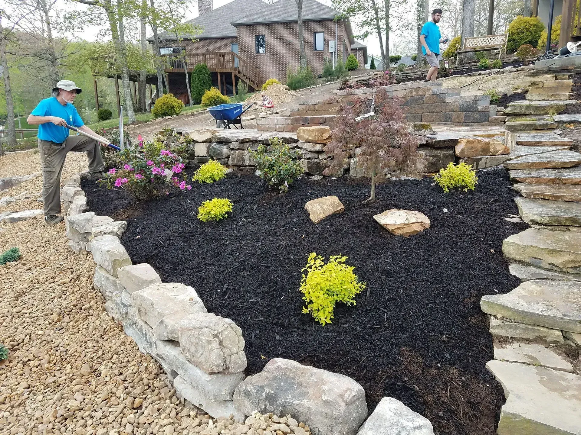 People landscaping a stone terrace garden with black mulch and small flowers.