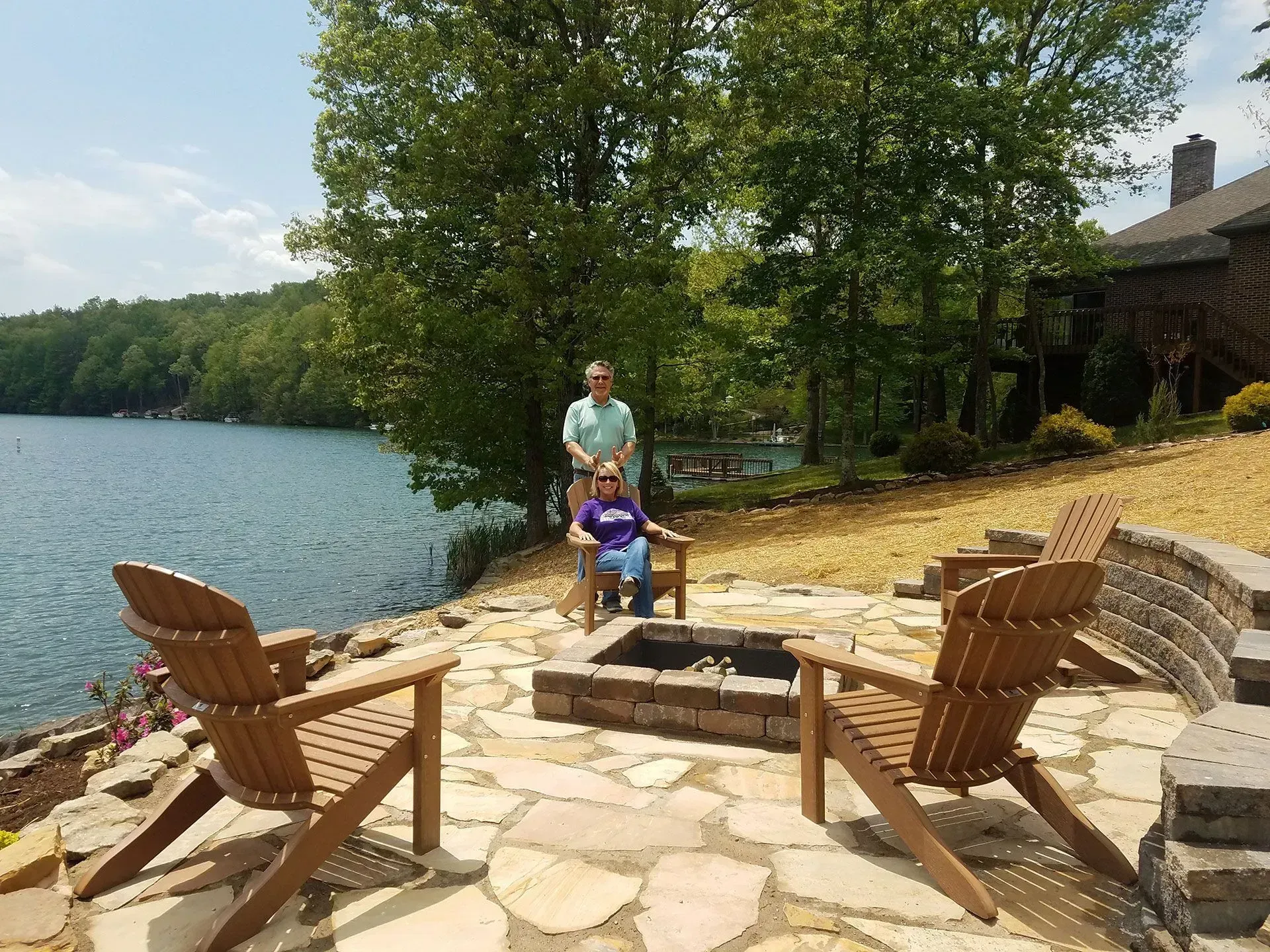 Lakeside patio with Adirondack chairs around a fire pit, a person seated under trees, and water in the background