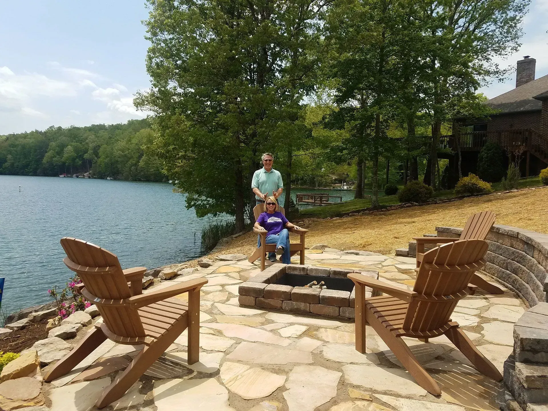 Person and child by lakeside fire pit with Adirondack chairs and trees