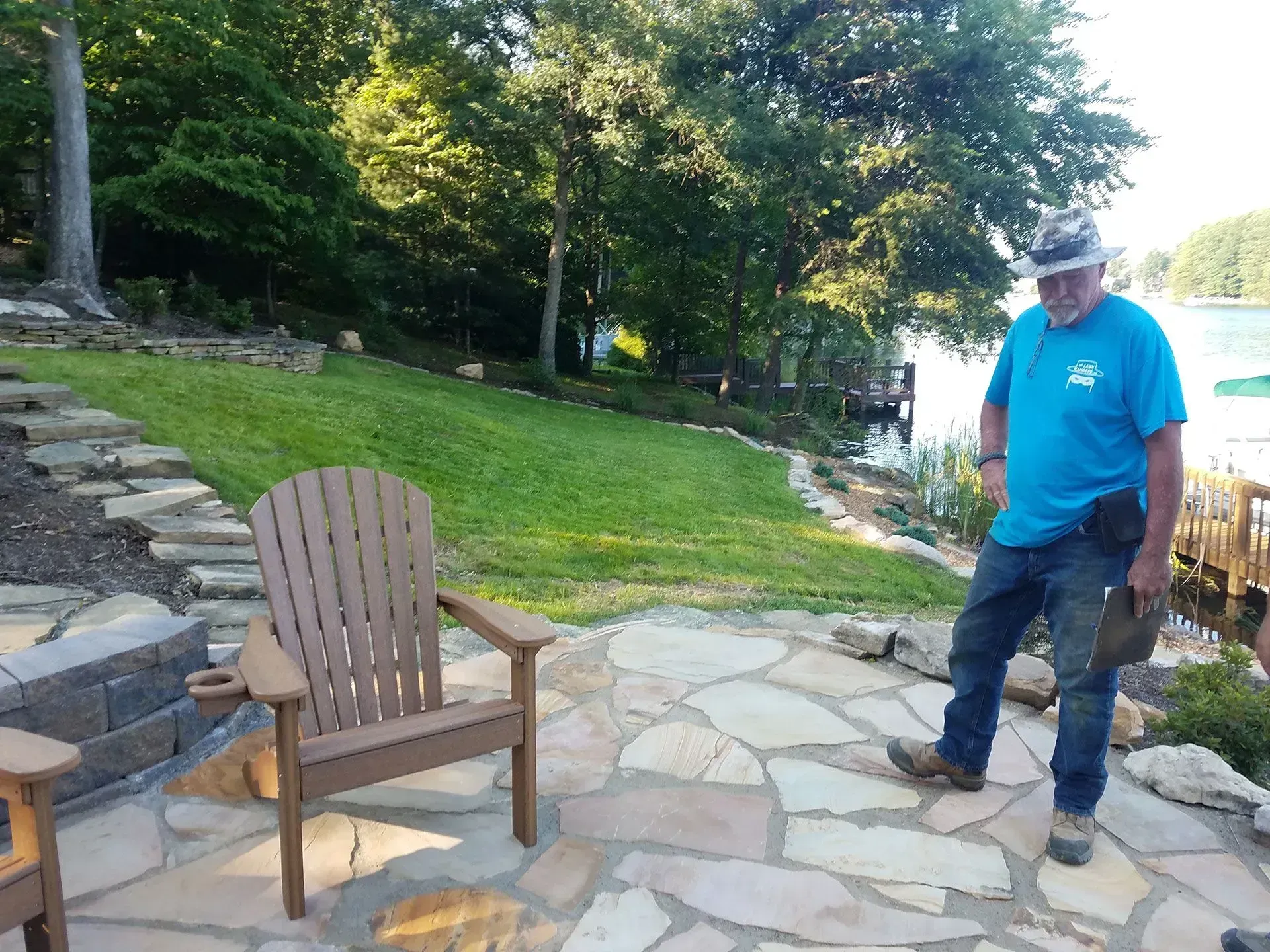 Man standing on a stone patio beside a wooden chair in a grassy yard near a lake