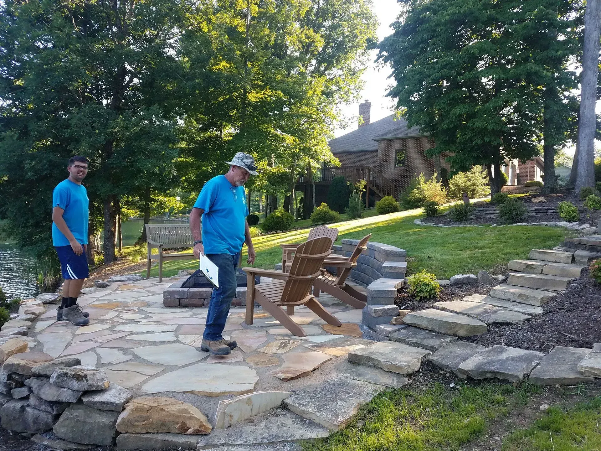 Two people in blue shirts moving wooden chairs on a stone patio in a garden