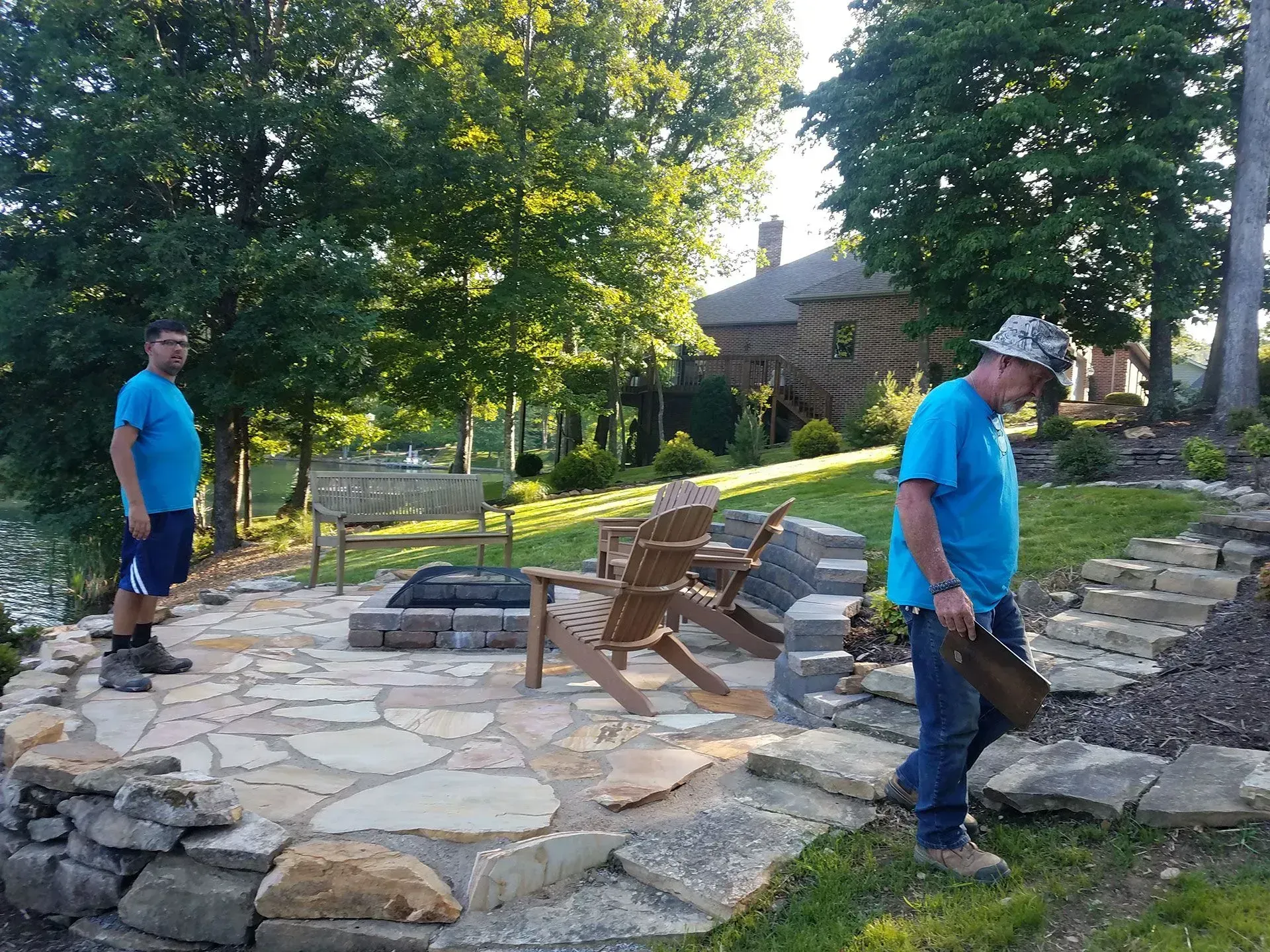 Two workers in blue shirts near a stone patio and fire pit in a wooded yard